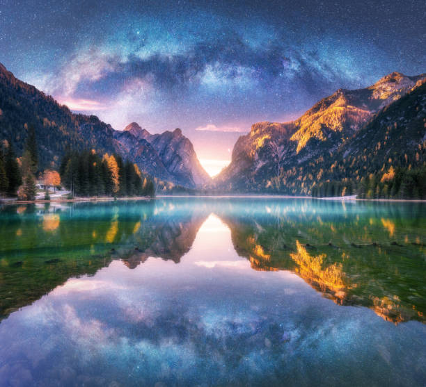 Photo of Milky Way reflected in water in mountain lake at starry night. Autumn landscape with purple sky with stars, blue fog, reflection, trees with colorful leaves, rocks in fall at sunset. Space and galaxy