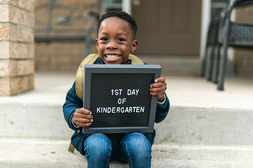 A cute boy of African America ethnicity sits on the patio stairs at his family home. He is holding a small black sign that reads first day of kindergarten. The boy is holding the sign on his lap. He is wearing a backpack and winter clothes.