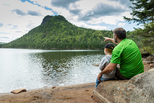 A father and his son are sitting on a rock and contemplating the lake and mountains during their summertime vacations.