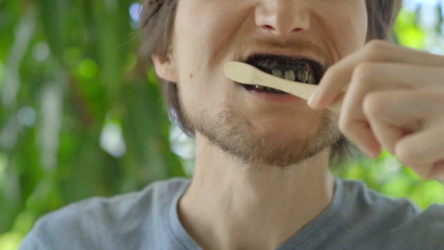 A young man in a green environment brushes his teeth with a black active charcoal powder for teeth whitening. He uses a toothbrush made of a bamboo. Concept of eco friendly zero waste bamboo products