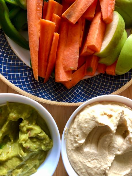 Image of hummus and guacomole platter, healthy vegan appetiser, hommus / houmous made from chickpeas, olive oil, tahini paste and garlic served with sliced carrot, green pepper and apple, elevated view Stock photo of houmous and guacomole platter, healthy eating vegan appetiser, Greek cuisine food (hommus / hummus) made from chickpeas, olive oil, tahini paste and garlic, served with green pepper, carrot and Granny Smith apple slices. appetiser stock pictures, royalty-free photos & images