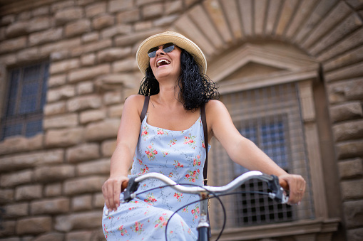 A beautiful woman rides a bicycle with a dress and a straw hat at the ancient streets of Florence Italy.