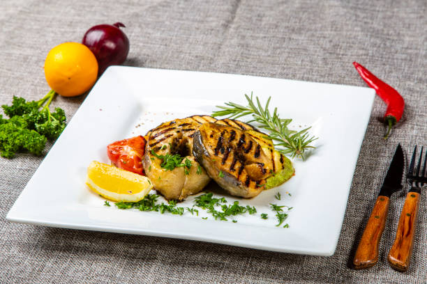 Sturgeon steak with vegetables. Delicious sturgeon steaks are grilled and served on a white square plate with lemon and tomato. The plate is on a gray canvas, next to the onion, lemon, red pepper, knife and fork. bistec de esturión con verduras. deliciosos filetes de esturión se asan a la parrilla y se sirven en un plato cuadrado blanco con limón y tomate. el plato está sobre un lienzo gris, junto a la cebolla, limón, pimiento rojo, cuchillo y tenedor