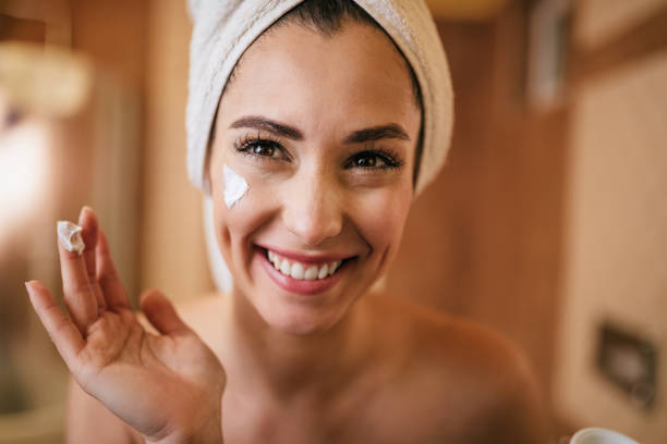 Young happy woman applying moisturizer on her face in the bathroom. Beautiful smiling woman applying face cream in the bathroom. woman wrapped in bath towel stock pictures, royalty-free photos & images