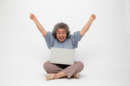 Excited senior asian woman feeling winner celebrating victory online business success and sitting on floor with laptop isolated on white background, Freelance mature concept