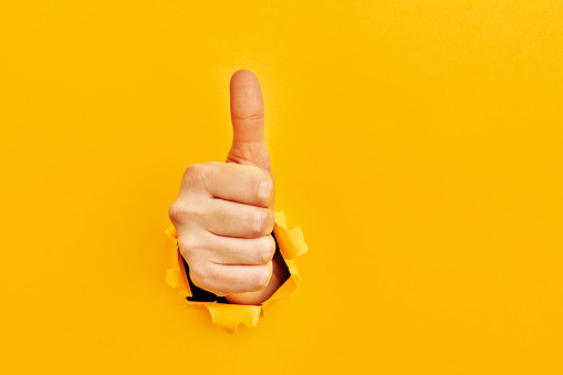 Human hand reaching through torn yellow paper sheet showing thumbs up