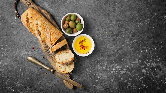 Top view of fresh wholegrain bread with green olive and oil in white bowls on wooden board and gray concrete background with copy space