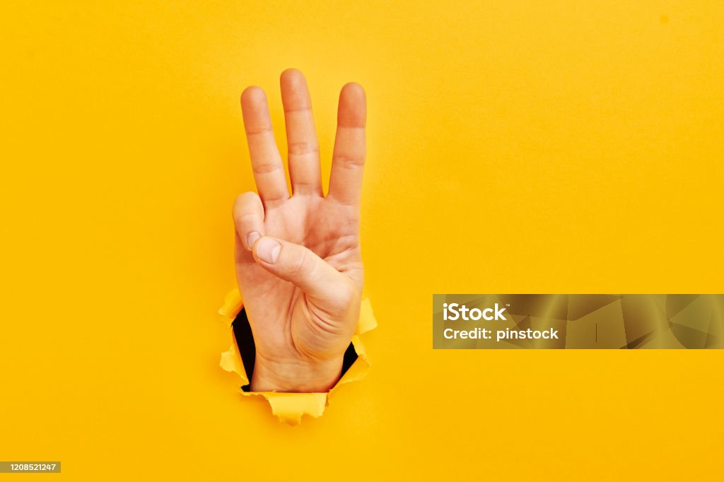 Human hand reaching through torn yellow paper sheet showing number three Number 3 Stock Photo