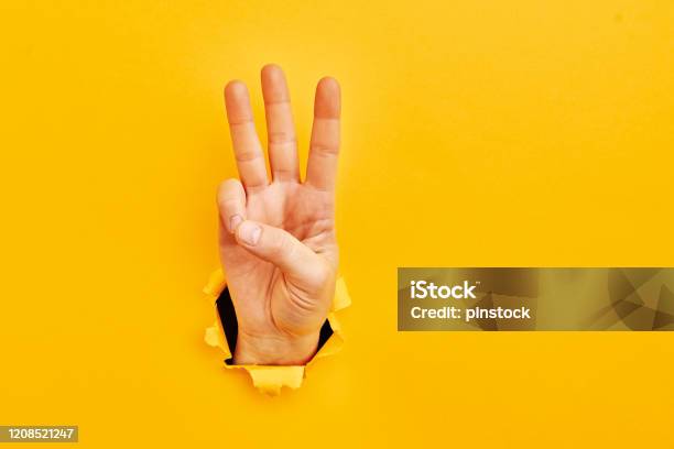 Human Hand Reaching Through Torn Yellow Paper Sheet Showing Number Three Stock Photo - Download Image Now