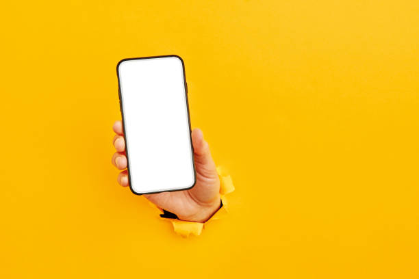 istock Human hand reaching through torn yellow paper sheet showing smart phone 1208521358
