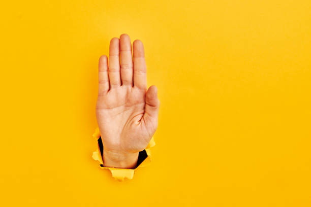 Human hand reaching through torn yellow paper sheet showing \