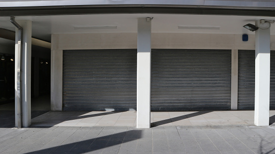 Shop closed with rolling shutters lowered under a porch with columns. White wall, shadows and in front a tiled pavement. Copy space