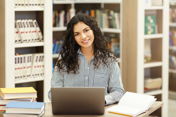 fille confiante de latina utilisant l’ordinateur portatif dans la bibliothèque moderne - library young adult bookstore people photos et images de collection