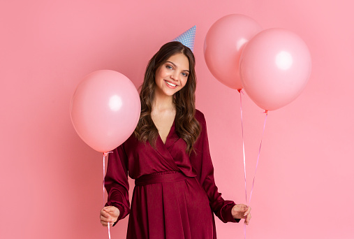 Birthday Girl. Portrait Of Attractive Young Woman With Helium Balloons And B'Day Hat Posing Over Pink Background With Free Space