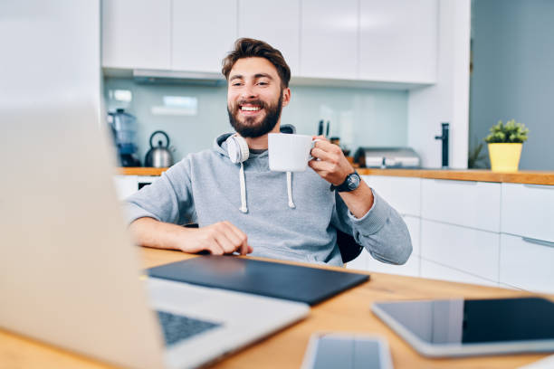 joven desarrollador web alegre bebiendo café mientras se toma descanso del trabajo en la oficina en casa - coffee break home interior cafe coffee fotografías e imágenes de stock