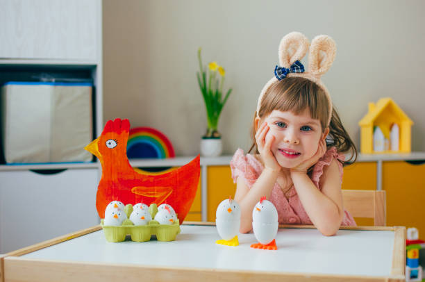 イースターの装飾で遊ぶバニーの耳を持つ笑顔の小さな女の子は、紙の鶏と鶏を塗った - bunny painting ストックフォトと画像