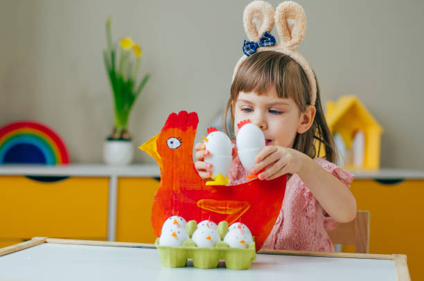 イースターの装飾で遊ぶバニーの耳を持つ笑顔の小さな女の子は、紙の鶏と鶏を塗った - bunny painting ストックフォトと画像