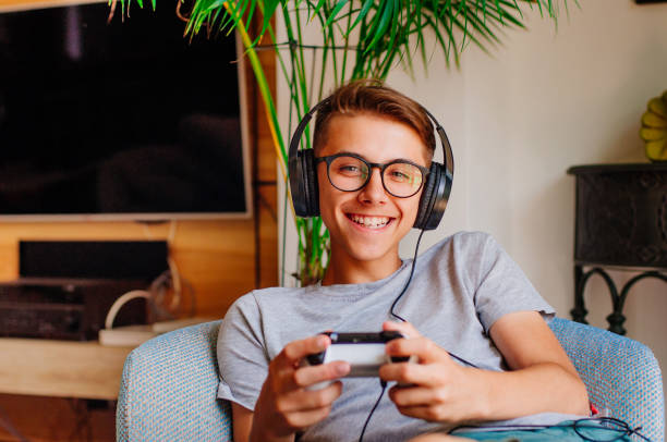 console de jeu de garçon d’adolescent de sourire avec des écouteurs s’asseyant dans la présidence à l’intérieur - teenager video game gamer child photos et images de collection