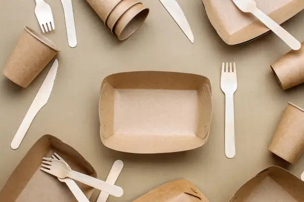 Disposable eco friendly food packaging. Brown kraft paper food containers, forks and knifes on beige background. Top view, flat lay.