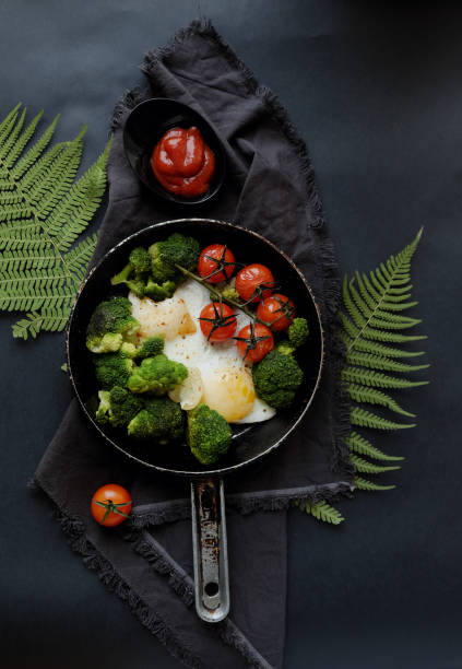 Beautiful healthy breakfast: broccoli, cherry tomatoes and fried eggs in a pan, which stands on a wooden stand on a dark background. Wooden cooking appliances.