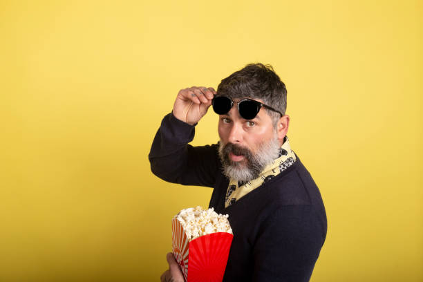 istock Handsome man with sunglasses in profile looking at camera holding a box full of popcorn on yellow background. 1208546853
