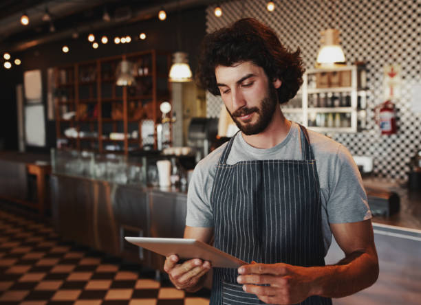 istock Young cafe owner checking for online order on digital tablet while standing in coffee house wearing black apron 1208546907