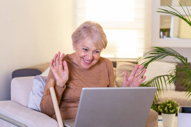 Happy mature woman waving to someone while having a video call over laptop at home. Gray-haired senior woman waving hand in front of laptop while having video call with her family members. mujer feliz madura saludando a alguien mientras tiene una videollamada sobre computadora portátil en casa. mujer mayor de pelo gris agitando la mano delante de la computadora portátil mientras tiene videollamada con los miembros de su familia.