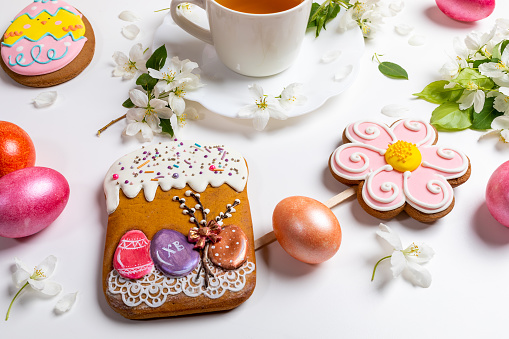 Close-up Easter festive gingerbreads as if Easter cake, flower and funny egg and colored eggs with cup of tea and white Apple tree twigs on white background. Easter morning tea time. Easter breakfast.