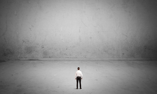 A tiny elegant businessman standing in large empty urban space with concrete walls and grey background concept