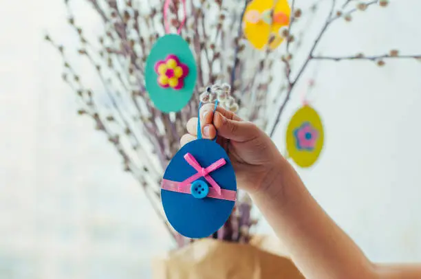 Close up of child hanging Easter eggs cutted from color paper decorated with ribbon and buttons on willow branches. Idea for DIY decoration with children.