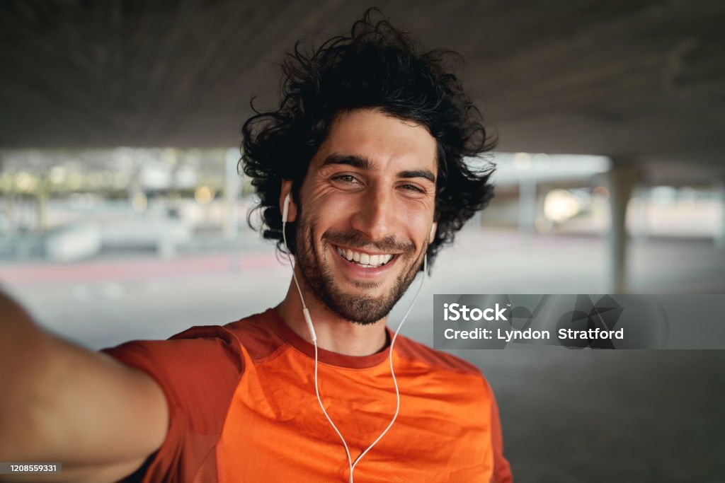 Portrait of a smiling fit young man with earphones in his ears taking selfie outdoors - pov shot of a man looking at the camera smiling taking a selfie Portrait of a happy sporty young man taking a selfie looking into the camera Selfie Stock Photo