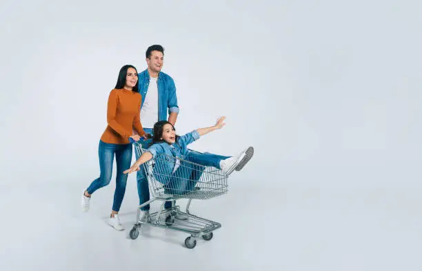 Full-length photo of a young man and his attractive wife, who are laughing, while riding their happy little child in a shopping cart.