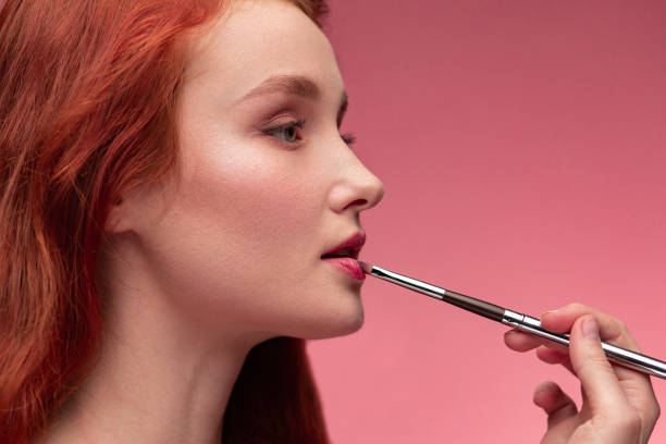 Makeup artist applies lipstick. Hand of make-up master, painting lips of young redhead girl. Portrait of beautiful healthy girl with pure clean skin and natural make-up. Beauty Concept Makeup artist applies lipstick. Hand of make-up master, painting lips of young redhead girl. Portrait of beautiful healthy girl with clean skin and natural make-up on pink background. Beauty Concept. rosy cheeks stock pictures, royalty-free photos & images