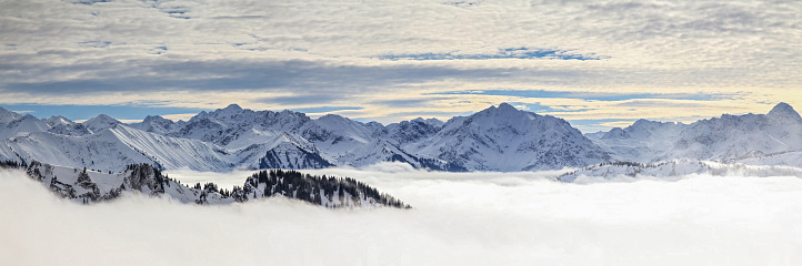 Steep mountain slopes and mountain ranges in low lying valley fog and trees shrouded in mist. Scenic snowy winter landscape in Alps, Allgäu, Bavaria, Germany. View from Riedberger Horn over Kleinwalsertal to the Allgauer Alps.
