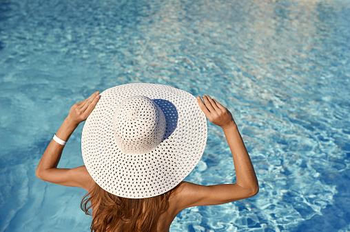 Rear view of woman in white hat sitting near pool on a sunny day. Sea travel concept with place for your text.