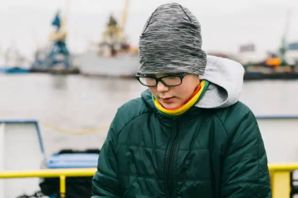 March 31, 2018 - Klaipeda, Lithuania: teenage caucasian boy standing in the ship port in cold weather and looking down concentrated