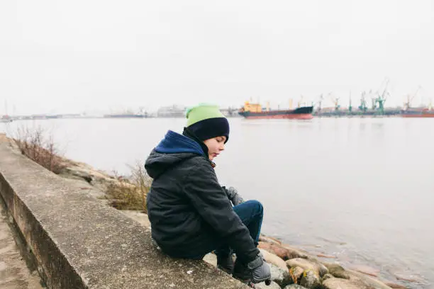 March 31, 2018 - Klaipeda, Lithuania: lonely child, a boy, sits and rests with his arm extended near sea on the rocks and looks down deep in his thoughts