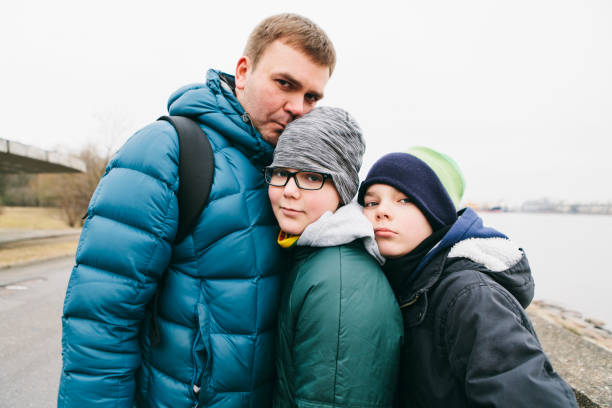 Handsome man - father - and his teenage sons standing close to each other, heads together, feeling love March 31, 2018 - Klaipeda, Lithuania: dad and two teenager boys pressed next to each other in a embrace while looking at camera, in their warm jackets and hats on a cold day lithuania photos stock pictures, royalty-free photos & images