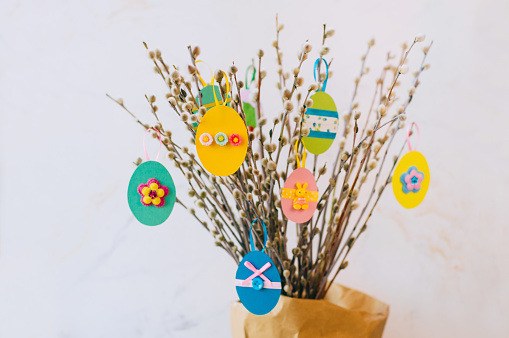 Easter eggs cutted from color paper decorated with ribbon and buttons on willow branches. Idea for DIY decoration with children. Selective focus on yellow paper egg.