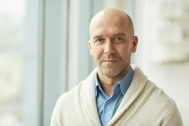 Balding Mature Man in Office Head and shoulders portrait of balding mature man wearing cardigan looking at camera while standing by window in white office, copy space completely bald stock pictures, royalty-free photos & images