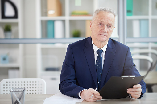 Portrait of successful senior businessman holding clipboard while posing at desk in office, copy space