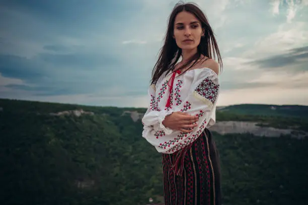 Photo of Atractive woman in traditional romanian costume on mountain green blurred background. Outdoor photo. Traditions and cultural diversity