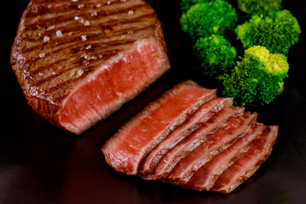 Sliced rare beef tenderloin with broccoli on black background.