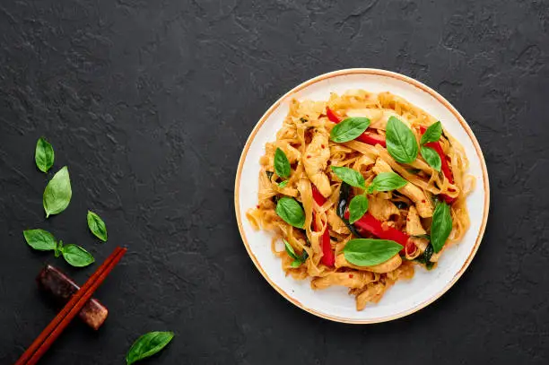 Thai Drunken Noodles or Pad Kee Mao in white plate at black slate background. Drunken Noodles is thai cuisine dish with Rice Noodles, Chicken meat, Basil, sauces and vegetables. Thai Food. Copy space.