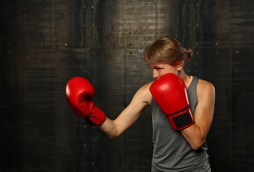Close up profile portrait of one mid adult athletic woman in sportswear over dark background, standing in boxing stance with hands in red gloves