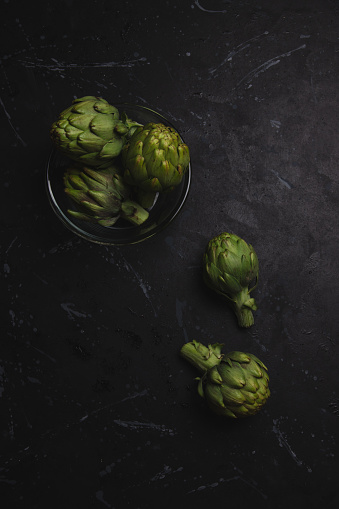 Artichokes in a glass bowl and more veggies on the table, on a dark distressed background