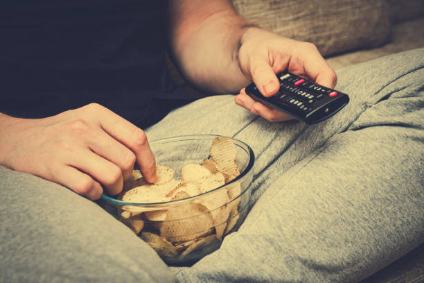 istock A man sits on a sofa eating chips from glassware and switches channels with a TV remote control. Toned. 1208604743