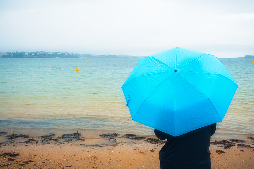 Back or rear view of a woman standing by the sea in rain coat and with a blue umbrella on a rainy and moody day. Weather concept with copy space. Orange and teal feel.