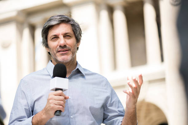 Journalist talking with a microphone outside a building while being filmed for television