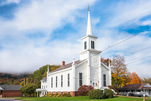Traditional white American wooden church at the foot of a forested hill covered in morning fog on a autumn day. Waterbury, VT, USA.